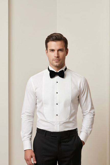 A man wearing a white long-sleeve tuxedo shirt with peak lapels, a black bow tie, and black trousers, standing against a light beige studio background.