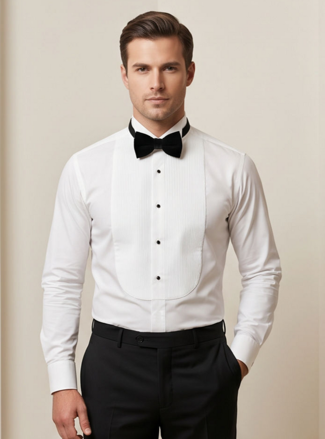 A man wearing a white tuxedo shirt with a black bow tie and black trousers, standing against a plain beige studio background.