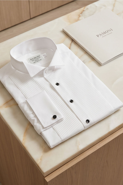 A folded white dress shirt with a subtle vertical texture and black buttons, displayed on a light marble surface next to a branded Pamoni booklet.