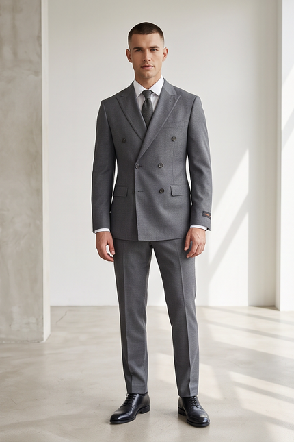A man wearing a gray double-breasted suit, white dress shirt, and gray tie, standing in a bright, modern room with concrete walls and large windows.